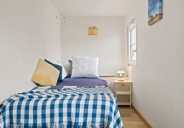 Un dormitorio con cama, almohadas, una colcha a cuadros, una mesita de noche con lámpara y una ventana.