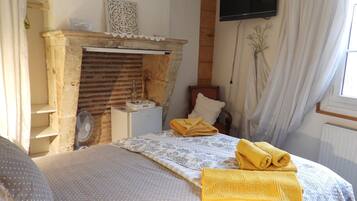 A bedroom with a brick fireplace, a bed with a polka dot bedspread, a wooden chair, a TV mounted on the wall, and a window with curtains.