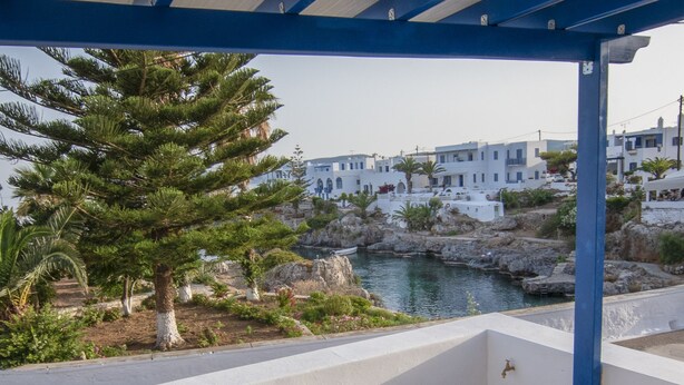 A covered outdoor seating area with a table set for two, overlooking a rocky coastline with buildings in the background. - Kanellis Studios (Kithira)