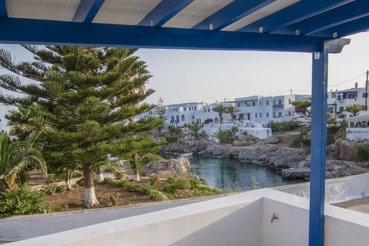 A covered outdoor seating area with a table set for two, overlooking a rocky coastline with buildings in the background.