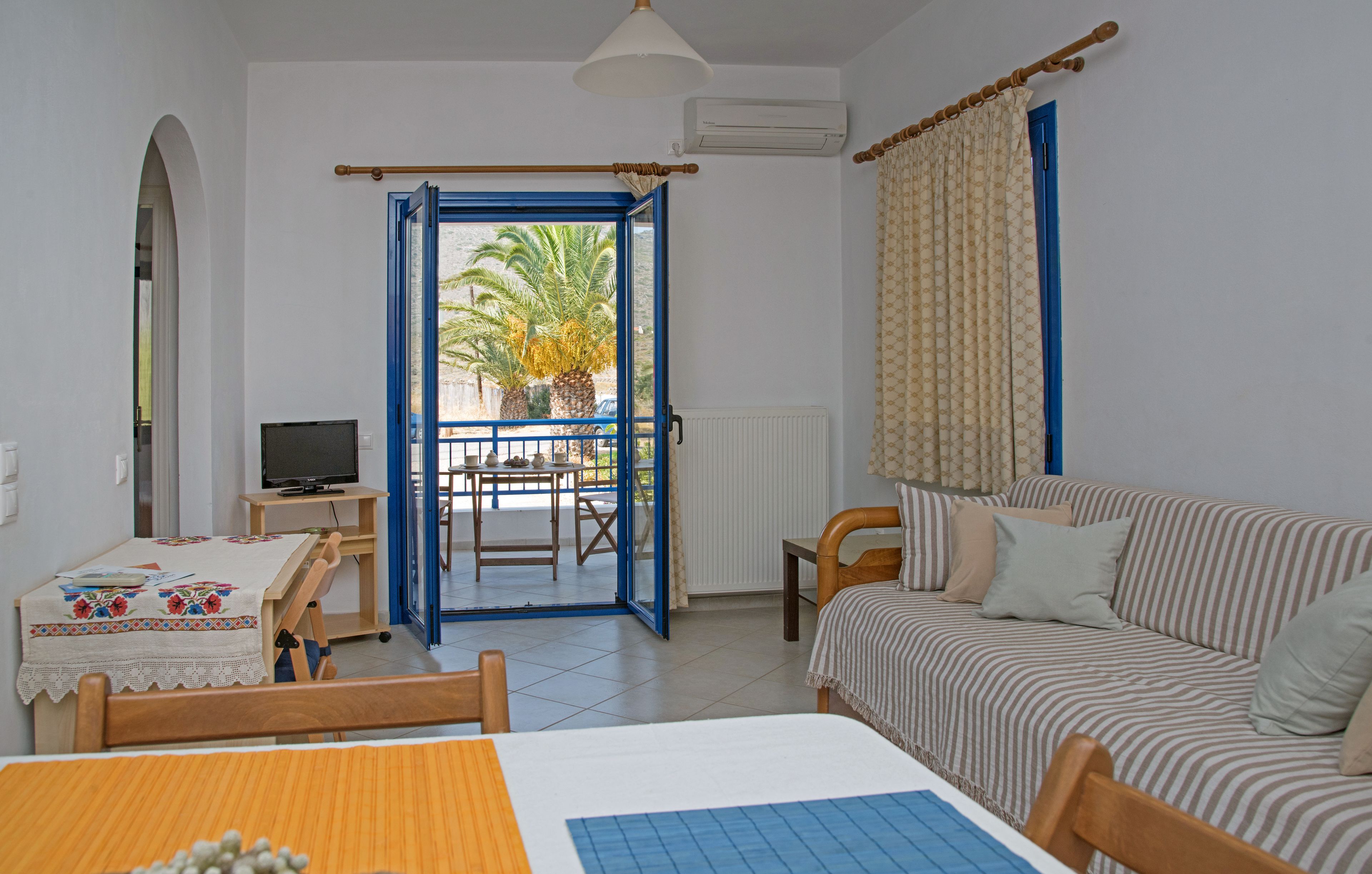 A room with a bed, a sofa, a TV, a table with a tablecloth, and a view of palm trees through an open door.