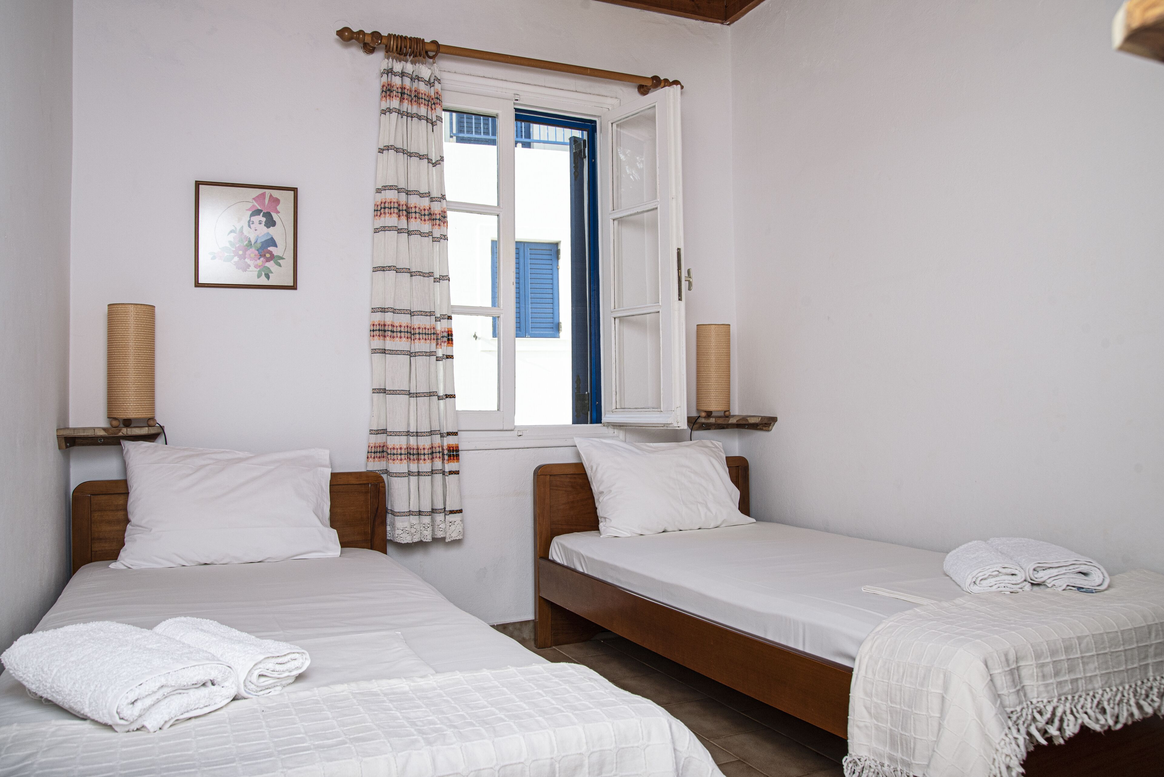 A bedroom with two single beds, a window with blue shutters, a curtain, and a framed picture on the wall.