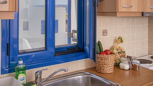 A kitchen with a blue-framed window, wooden cabinets, a stainless steel sink, and various kitchen items on the counter. - Kanellis Studios (Kithira)