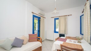 A room with a sofa, a dining table, and chairs. There are curtains on the windows, and an air conditioning unit is mounted on the wall. - Kanellis Studios (Kithira)