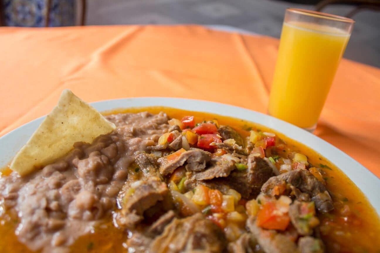 Um prato de ensopado de carne com feijão e legumes, uma tortilla e um copo de suco de laranja sobre uma toalha de mesa laranja.
