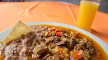 Um prato de ensopado de carne com feijão e legumes, uma tortilla e um copo de suco de laranja sobre uma toalha de mesa laranja.