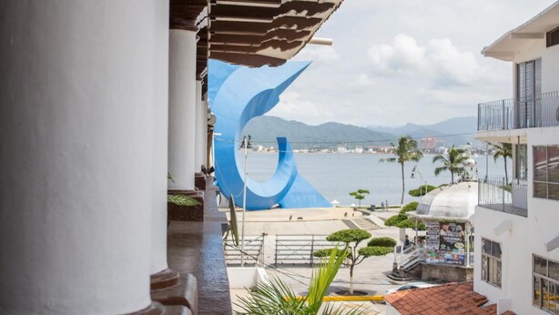 A view from a balcony overlooking a body of water with mountains in the distance, a large blue sculpture, and a building with a tiled roof. - Hotel Colonial (Manzanillo)