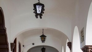A corridor with white walls, wooden door frames, and a tiled floor. There are two hanging lanterns and potted plants along the sides. - Hotel Colonial (Manzanillo)
