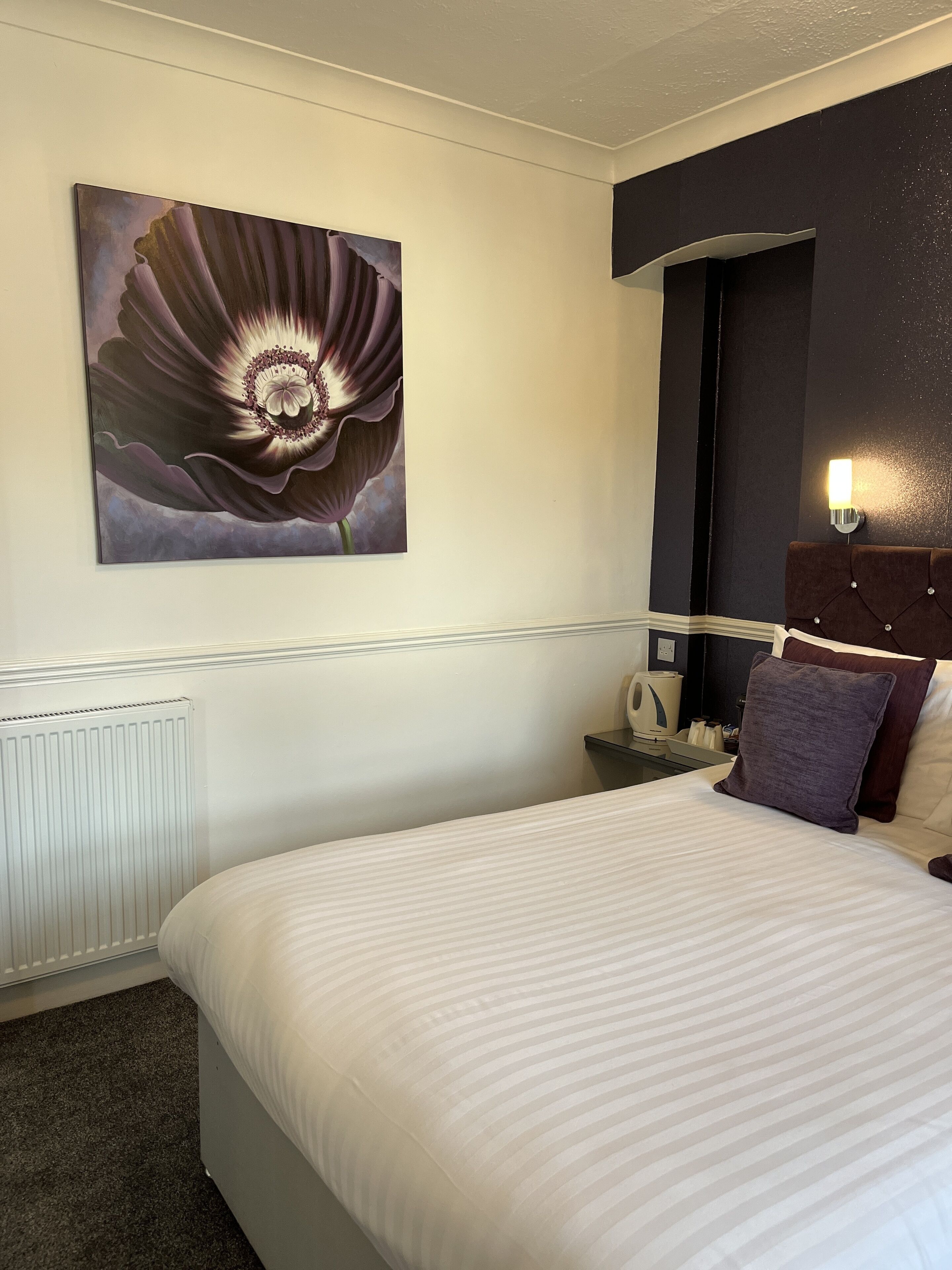 Ein Hotelzimmer mit einem großen Bett, einem Nachttisch, einer Wandleuchte und einem Blumenbild.