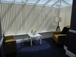 A conservatory with a blue carpet, a white table with a vase of flowers, yellow armchairs, and a floor lamp.