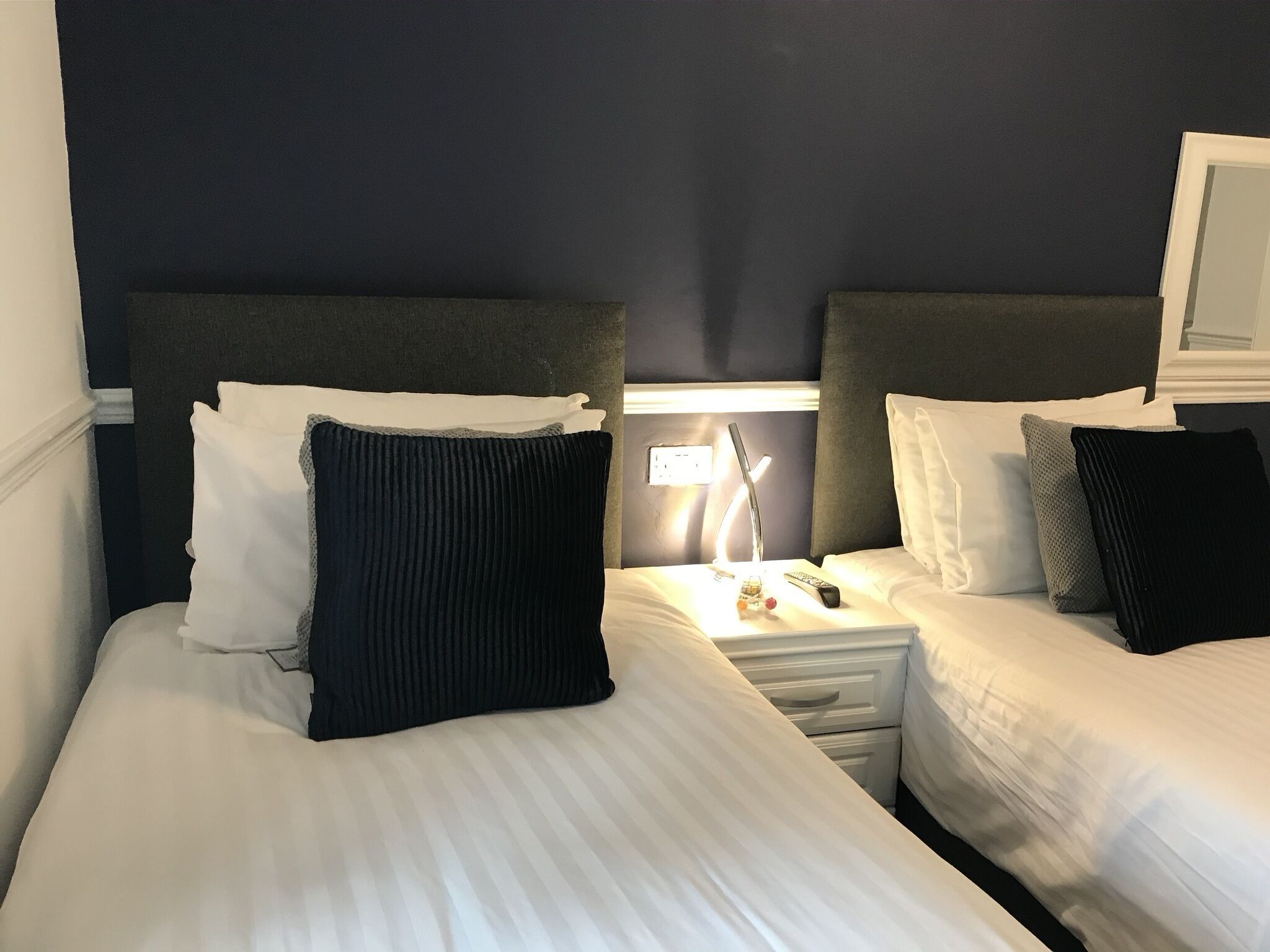 A hotel room with two beds, each with white linens and dark headboards. There is a nightstand with a lamp and a remote control between the beds.