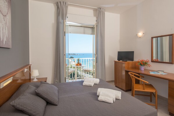 A hotel room with a bed, a desk, a chair, a TV, and a balcony with a view of the sea. - Hotel President Sea Palace (Noto)