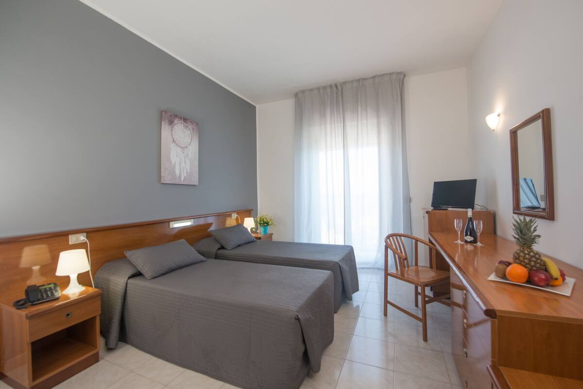 A hotel room with two beds, a desk with a computer and a fruit bowl, a chair, a small table with a lamp, and a painting on the wall.