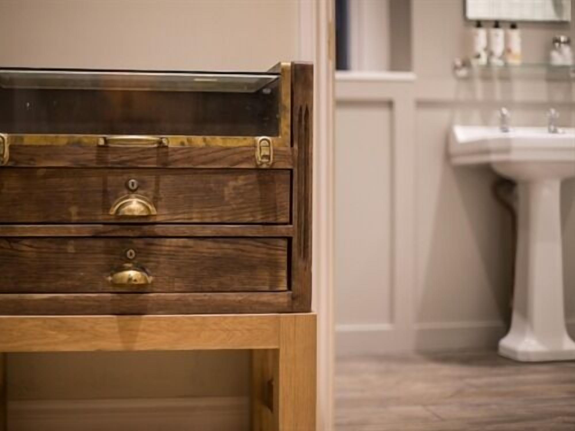 A wooden chest with brass handles and a drawer, placed in a room with a white pedestal sink and a mirror above it.