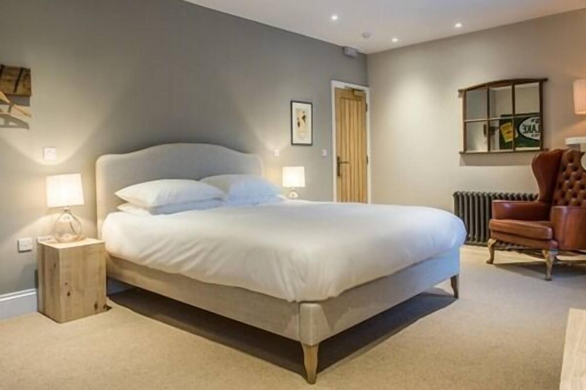 Un dormitorio con una cama grande, lámparas de noche, una mesita de madera, un sillón de cuero marrón y un radiador.