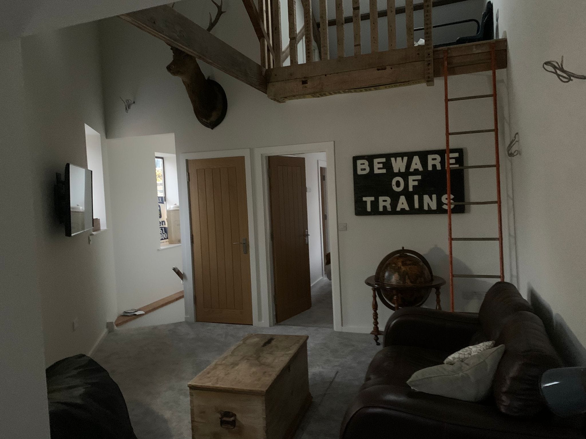 A living room with a wooden bench, a leather couch, a wall-mounted TV, and a 'Beware of Trains' sign.