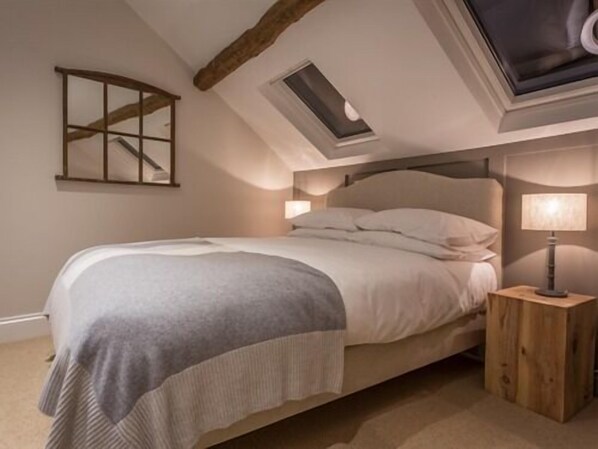 Un dormitorio con techo inclinado, tragaluz, cama con cabecera, mesita de noche con lámpara y suelo de madera.
