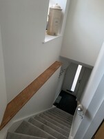 Una escalera con barandilla de madera, un estante pequeño con un recipiente blanco y una puerta con lámpara.