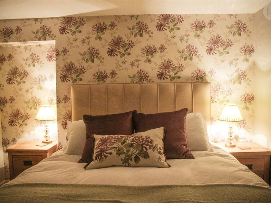 A bedroom with a floral wallpaper, a wooden headboard, two bedside lamps, and a bed with multiple pillows.