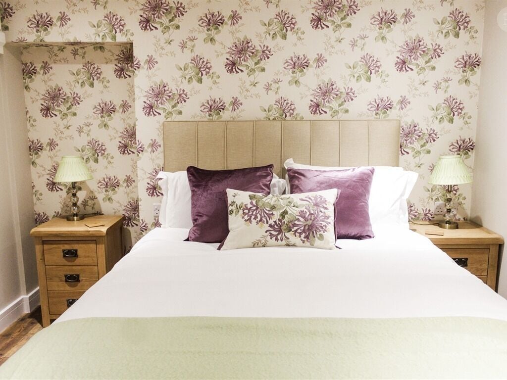 Una cama bien hecha con un cabecero estampado con motivos florales, dos mesitas de noche con lámparas y una mesita auxiliar de madera.