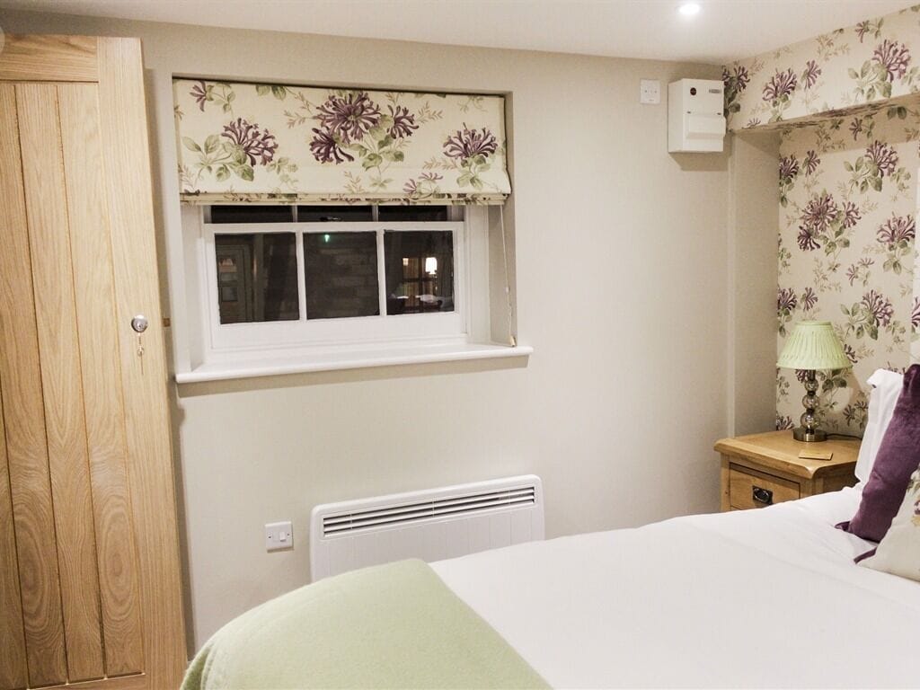 A bedroom with a bed, a window with floral curtains, a radiator, a wooden wardrobe, and a nightstand with a lamp.