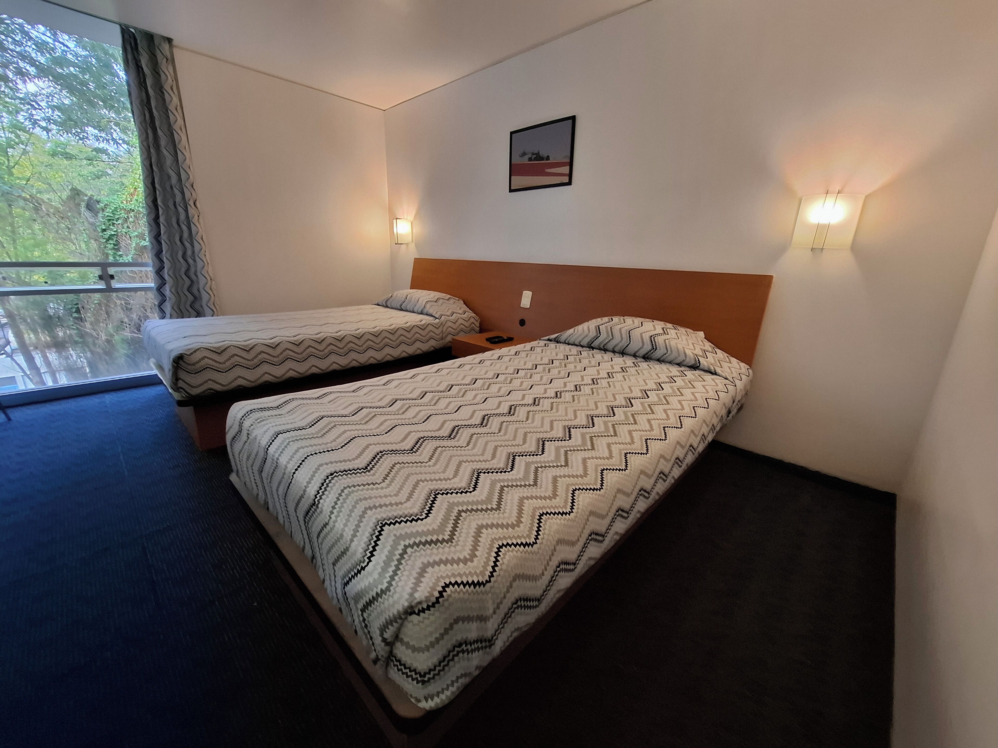 A hotel room with two beds, a window with curtains, a wall-mounted picture, and two wall sconces.