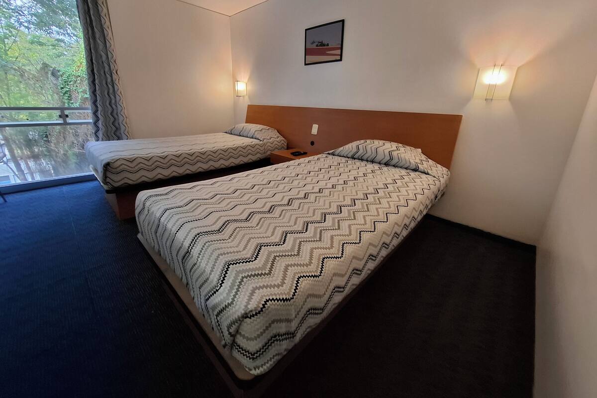 A hotel room with two beds, a window with curtains, a wall-mounted picture, and two wall sconces.