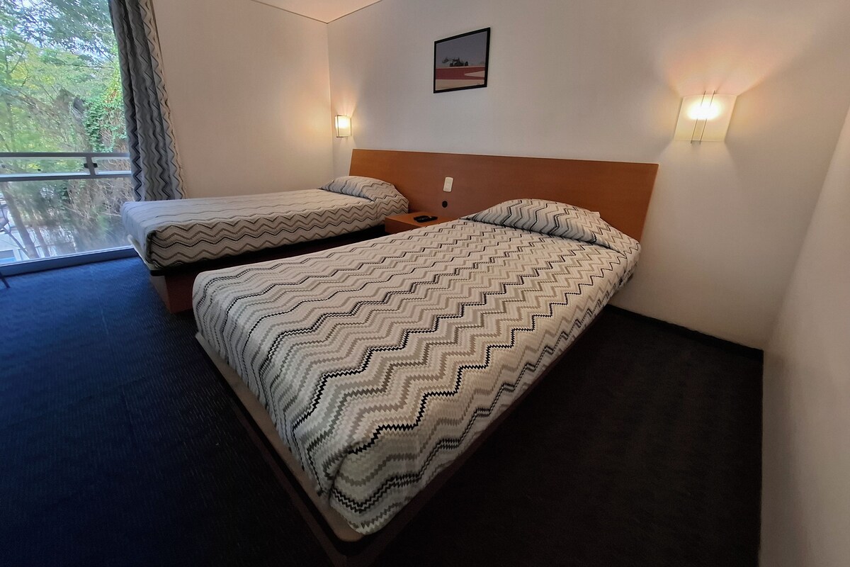 A hotel room with two beds, a window with curtains, a wall-mounted picture, and two wall sconces.