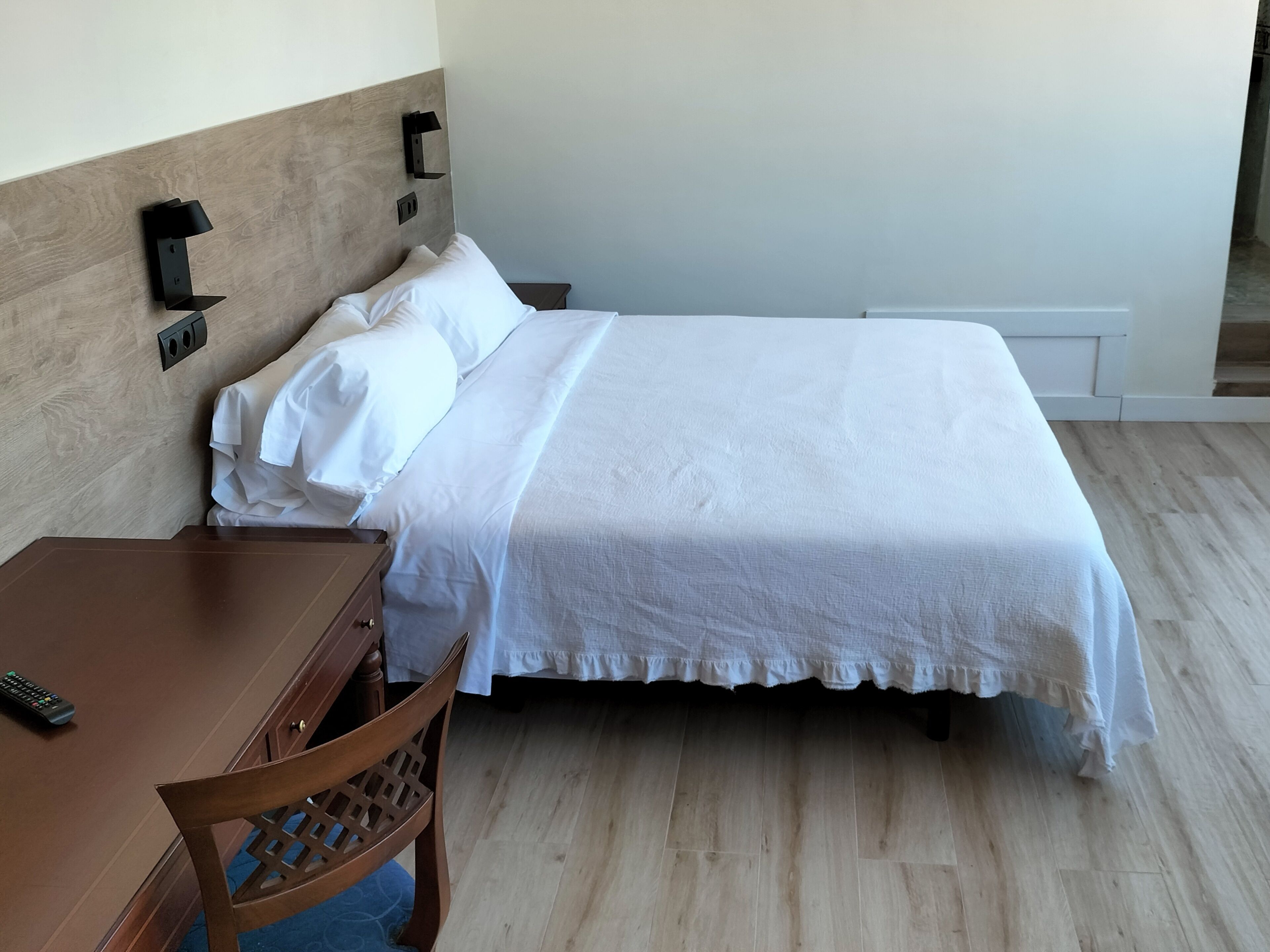 Un lit bien fait, avec du linge de lit blanc, une table de chevet en bois sur laquelle repose une télécommande, et un luminaire fixé au mur.