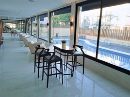 Un espacio interior moderno con vistas a una piscina, que cuenta con una hilera larga de taburetes y sofás, y una mesa de vidrio con una planta en maceta.