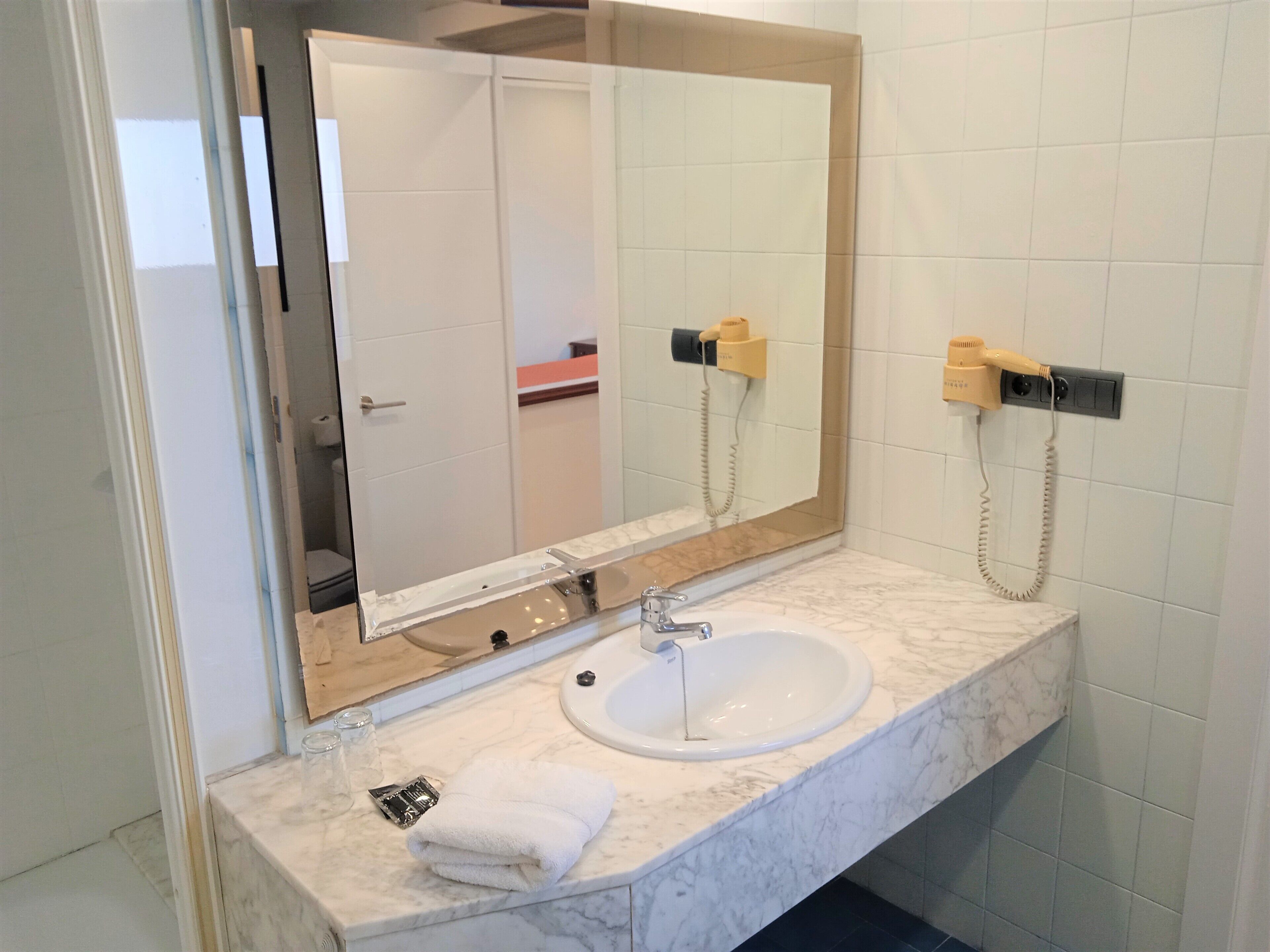 Een badkamer met een grote spiegel, een wastafel en een aan de muur bevestigde haardroger.