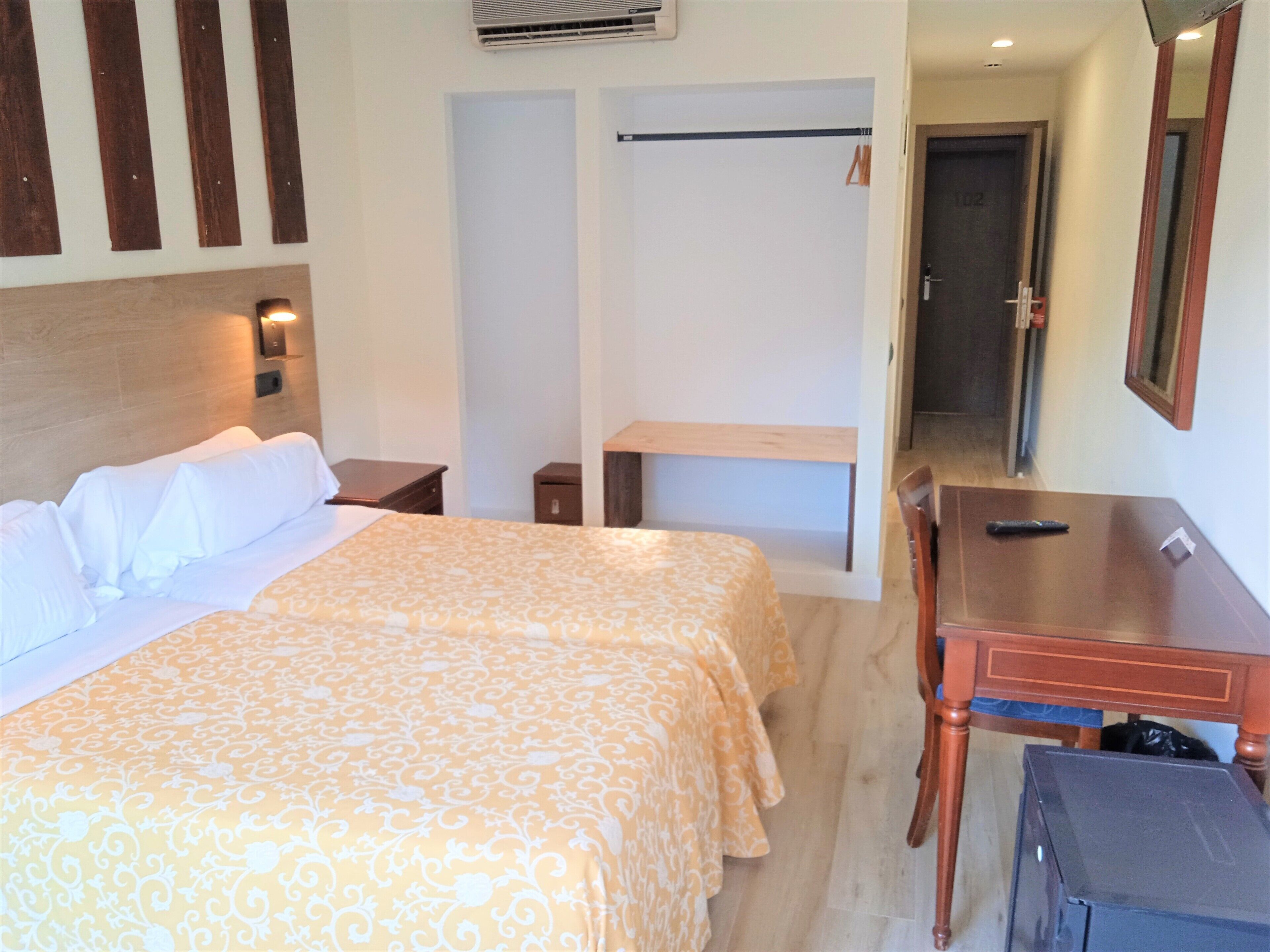 A hotel room with a large bed, a desk, a chair, a small table, a mirror, and a wardrobe.
