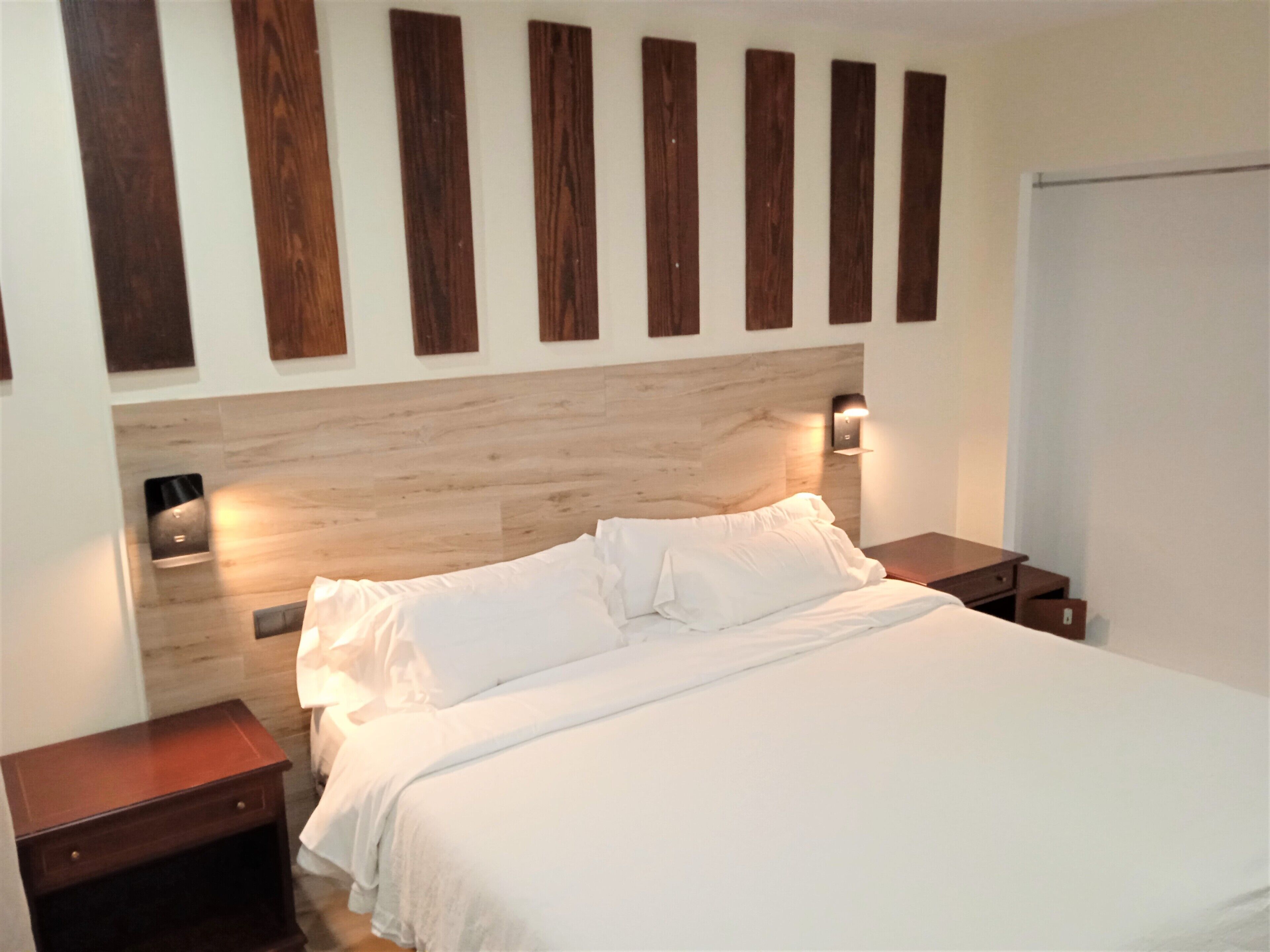 A neatly made bed with white linens, a wooden headboard, and two bedside tables with wall-mounted lamps.