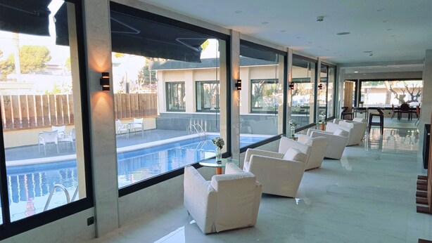 A modern indoor pool area with a clear view of the outside, white lounge chairs, and a glass-enclosed patio with outdoor seating. - Hotel 153 (Castelldefels)