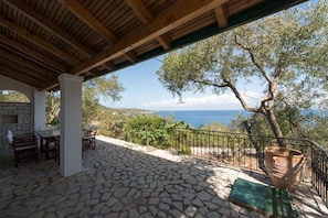 A covered outdoor area with stone flooring, a table with chairs, and a view of the sea. - Kipos Villas (Paxos)
