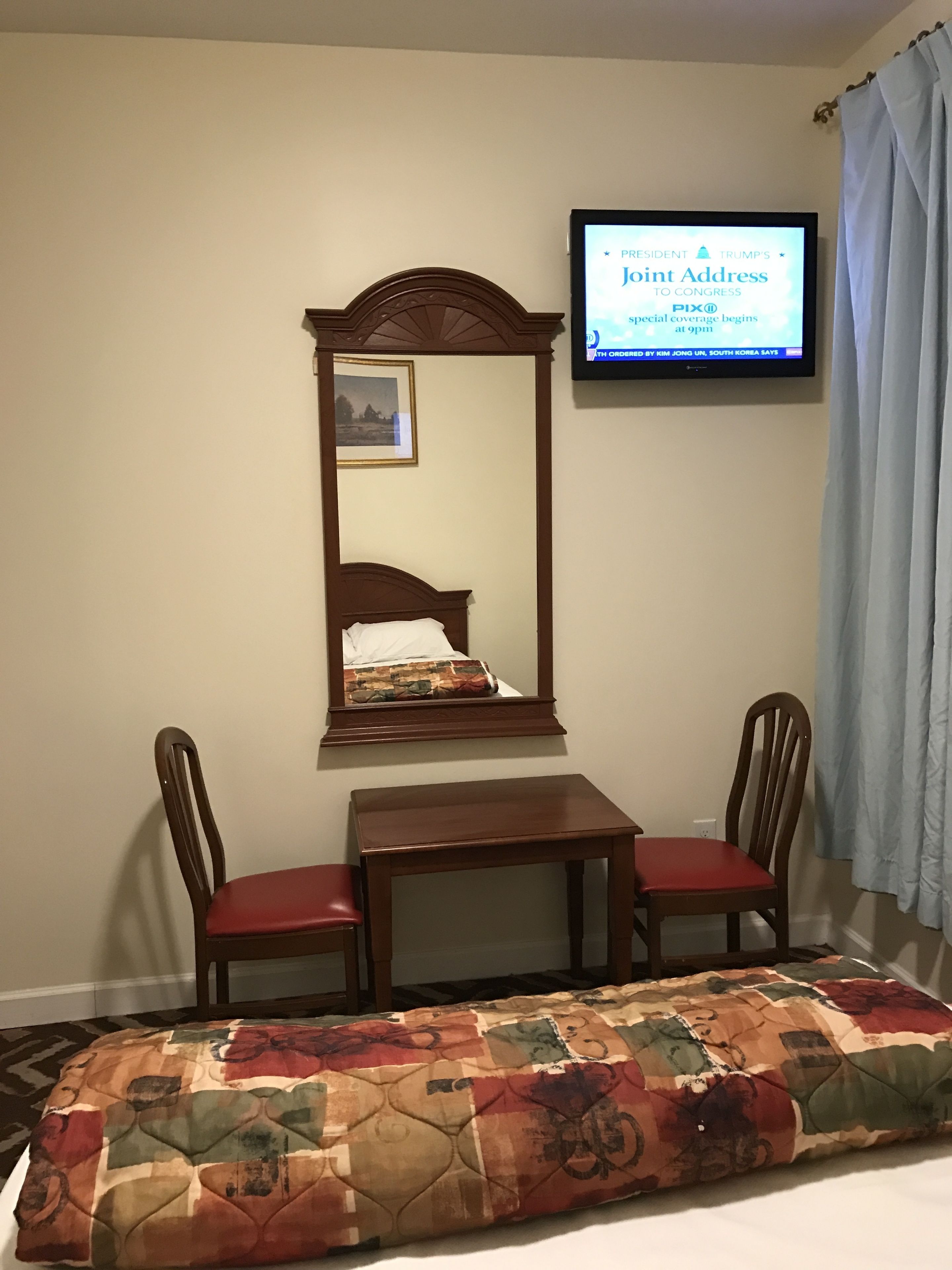 A hotel room with a wooden bed, a small wooden table, two wooden chairs, a television mounted on the wall, and a floral-patterned bedspread.