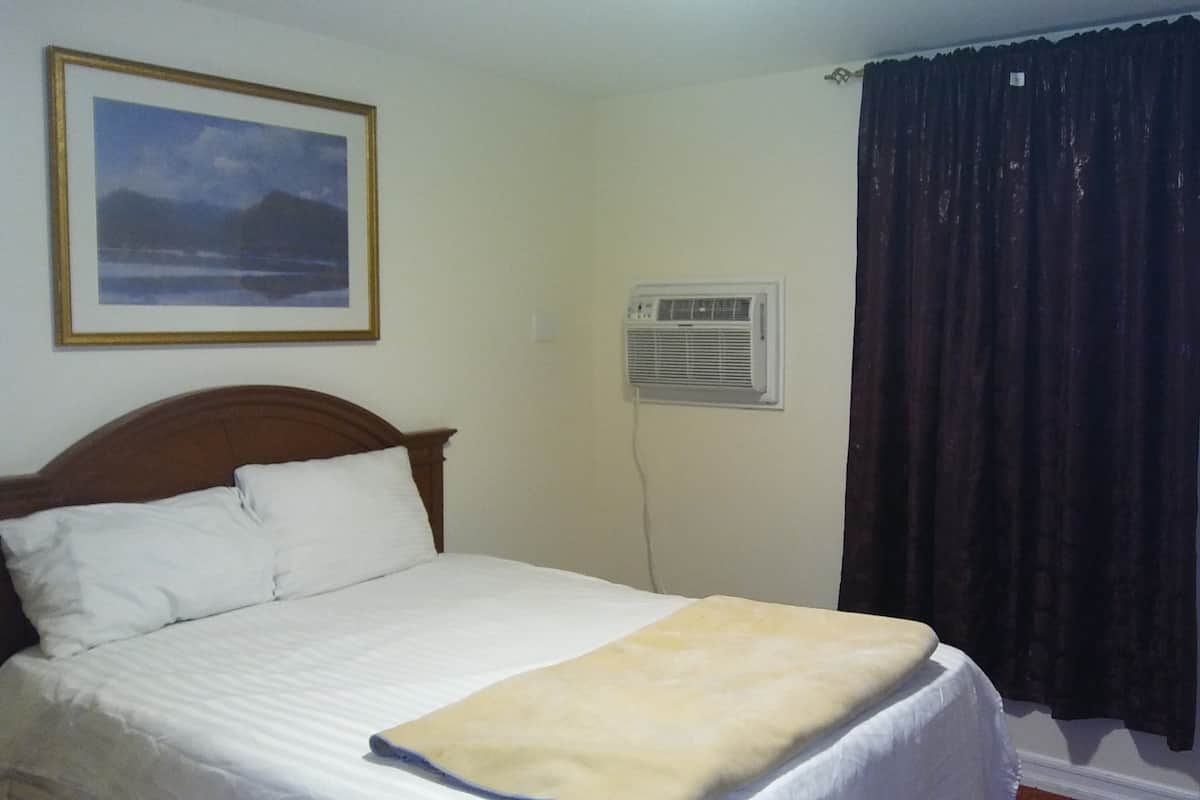 A bedroom with a bed, white bedding, a framed picture on the wall, an air conditioning unit, and a dark curtain.