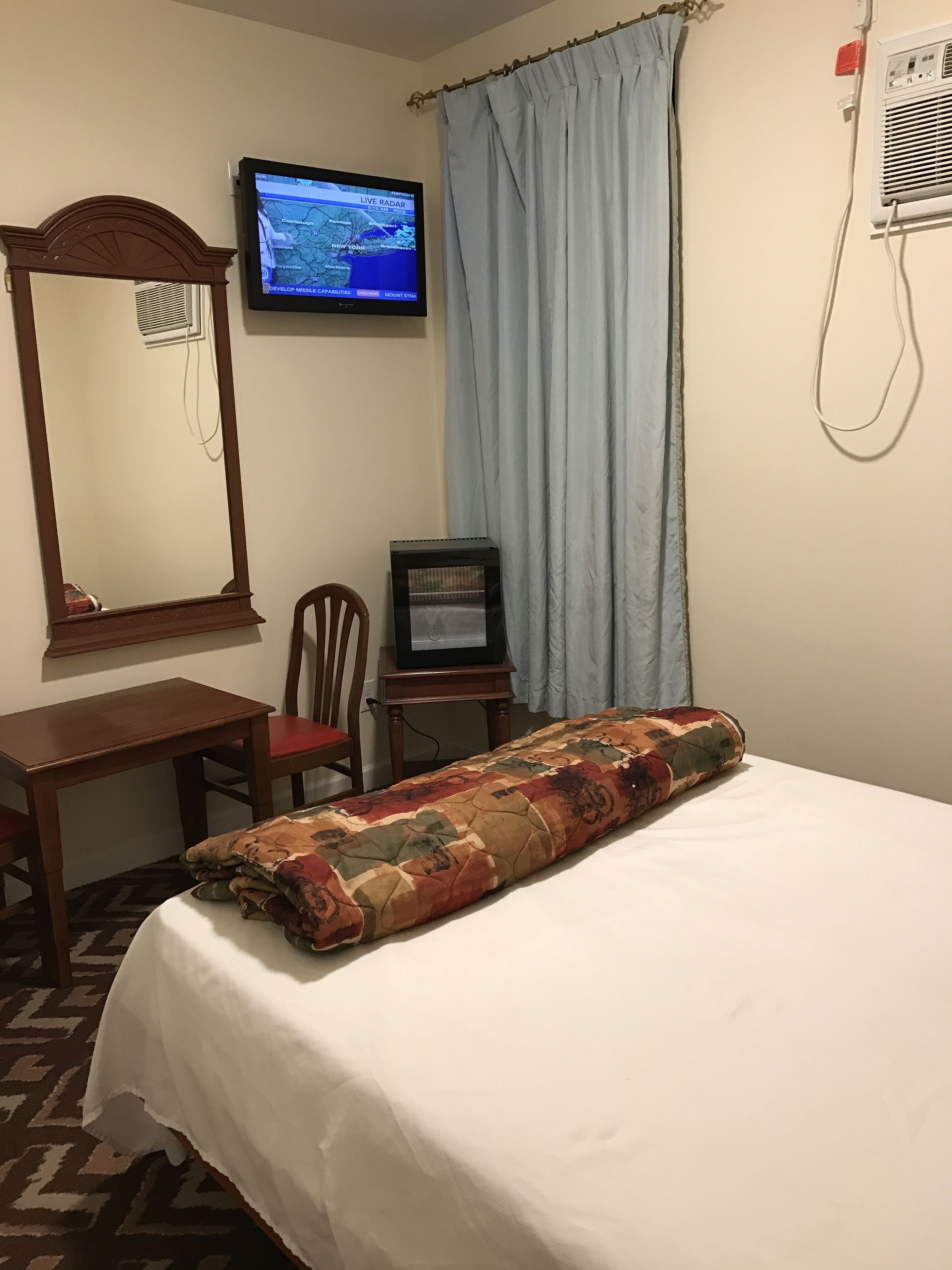 Quarto de hotel com cama, escrivaninha, cadeira, televisão e janela com cortinas.