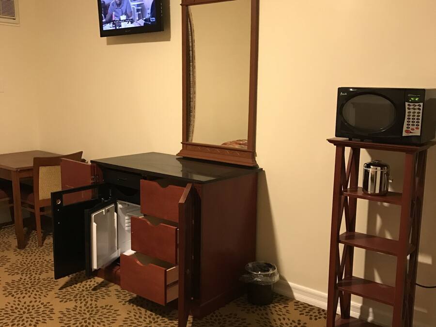 A hotel room with a wooden dresser, a television mounted on the wall, a microwave on a small table, and a patterned carpet.