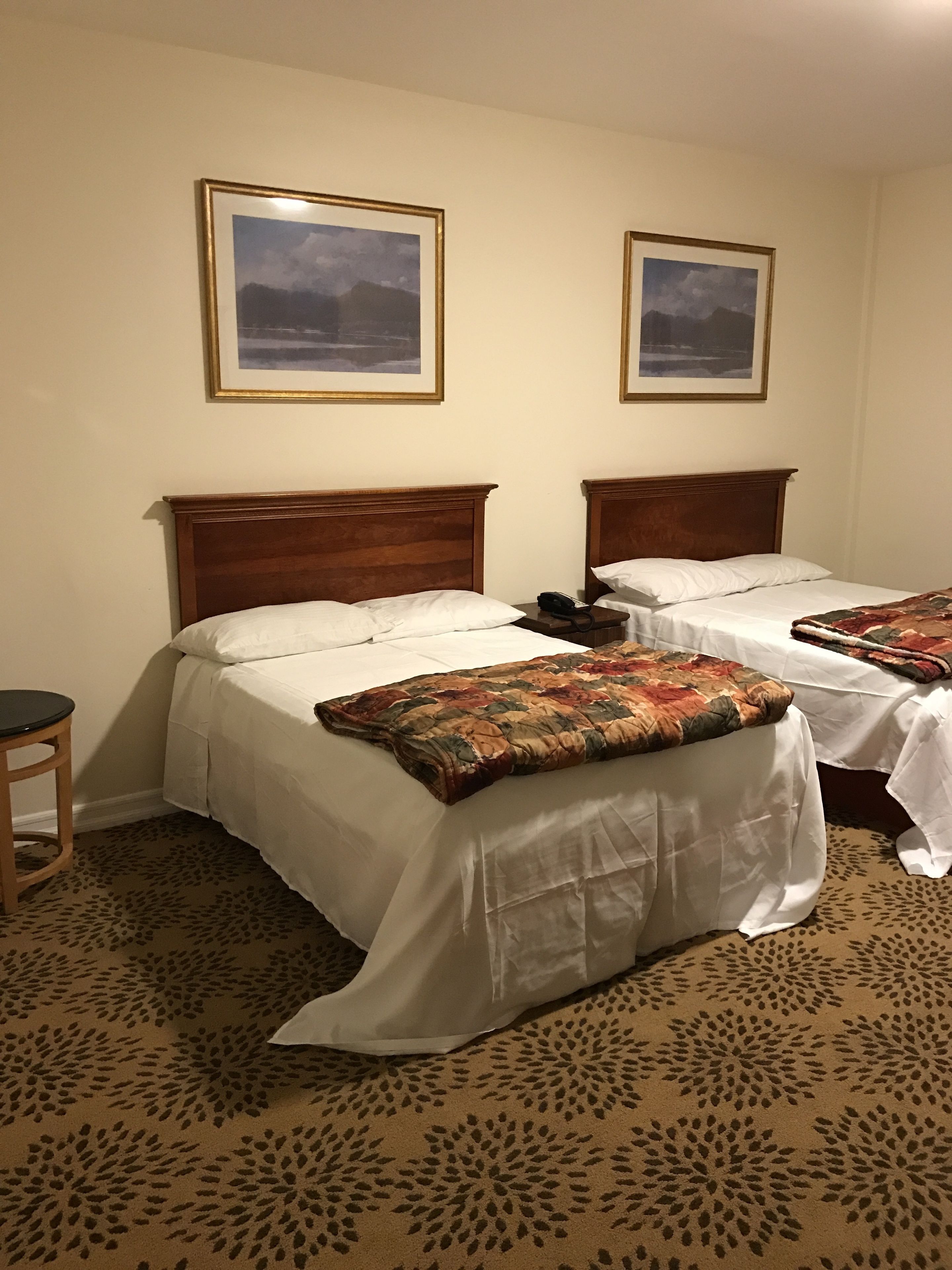 A hotel room with two beds, each with a wooden headboard, white bedding, and a patterned throw blanket. There is a small table with a chair, and two framed pictures on the wall.