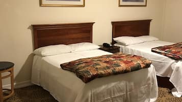 A hotel room with two beds, each with a wooden headboard, white bedding, and a patterned throw blanket. There is a small table with a chair, and two framed pictures on the wall.
