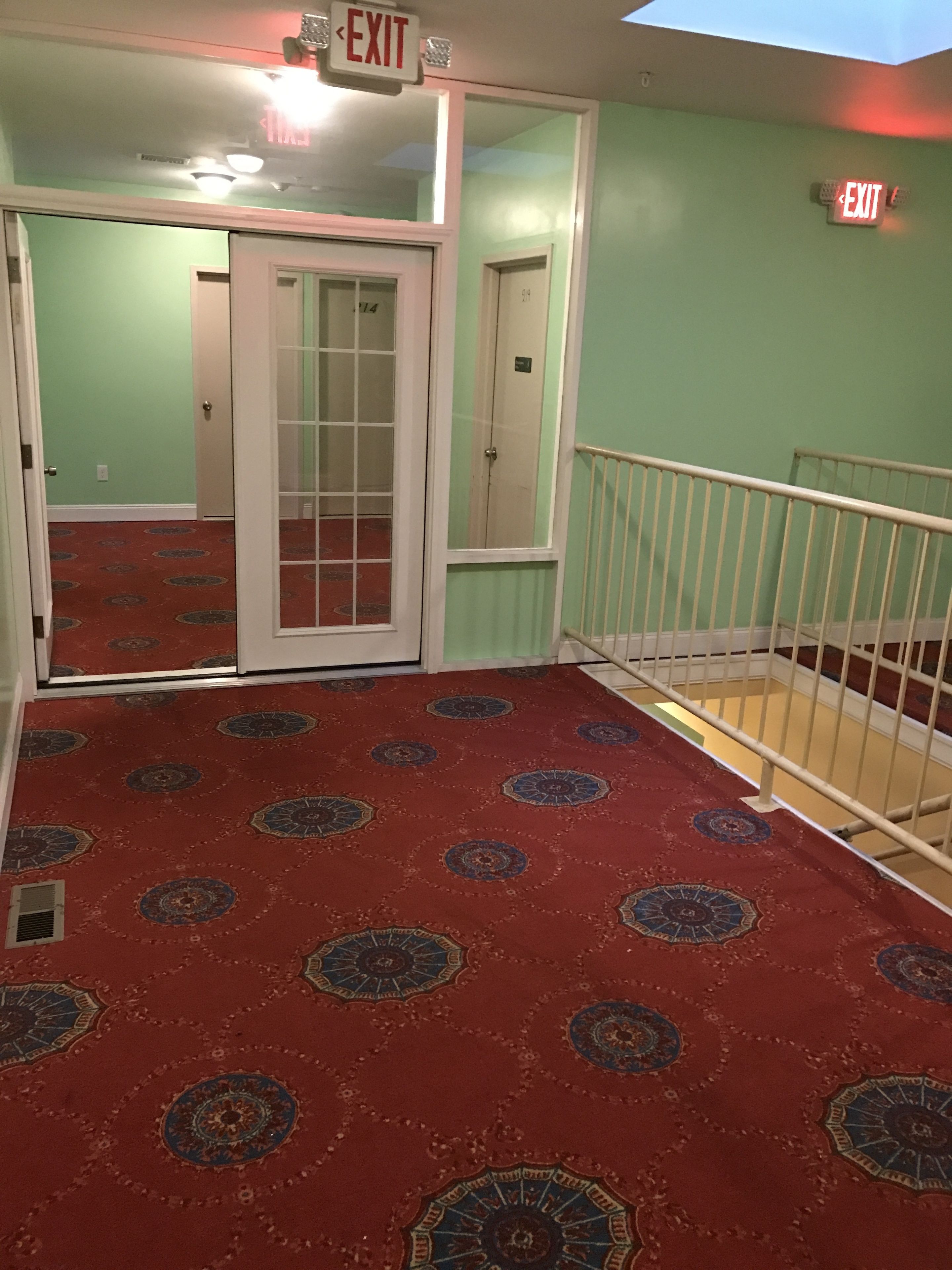 A hallway with patterned carpet, an exit sign, and a door leading to another room.