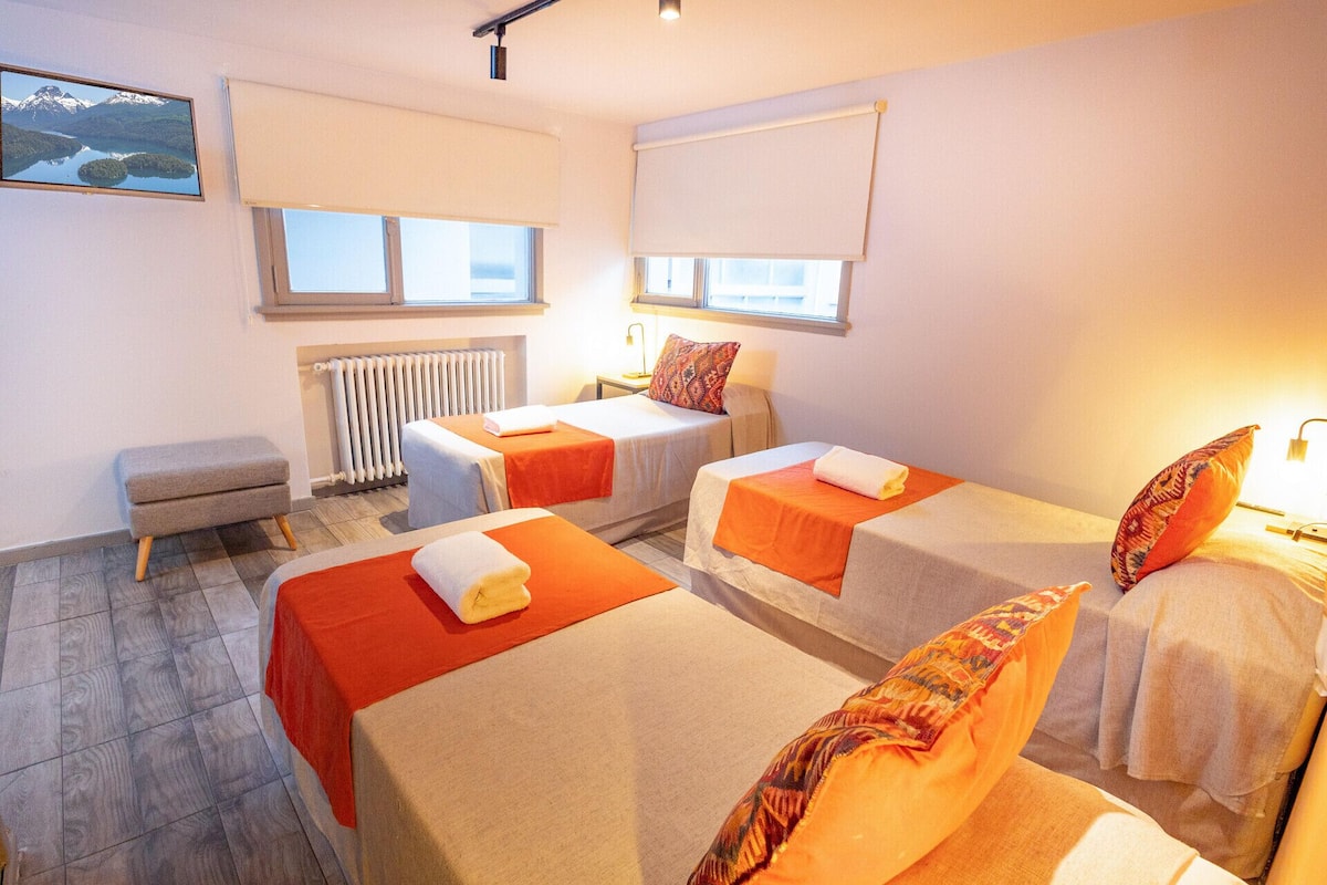 A room with two beds, a small bench, a radiator, and a window with blinds.