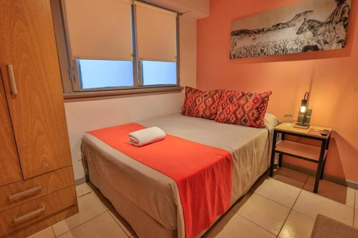 A bedroom with a bed, a nightstand, a window, and a wall-mounted artwork.