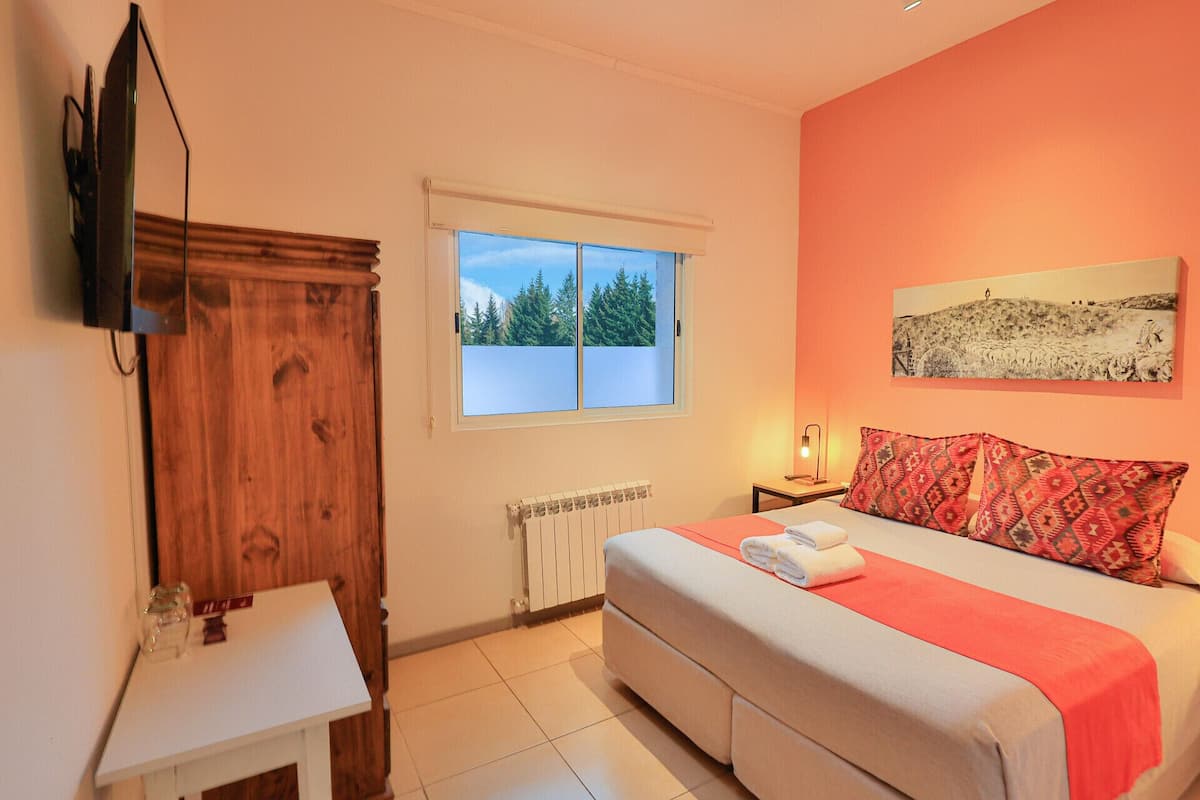 A bedroom with a bed, a window with a view of trees, a radiator, a bedside table, and a wall-mounted TV.