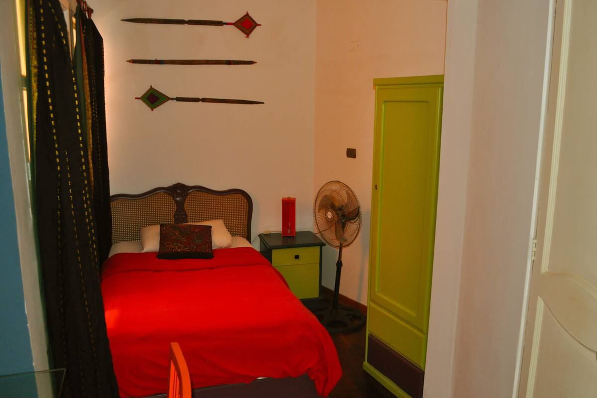 Un dormitorio con una colcha roja, un cabecero de madera, una mesita de noche con un ventilador y una puerta verde.