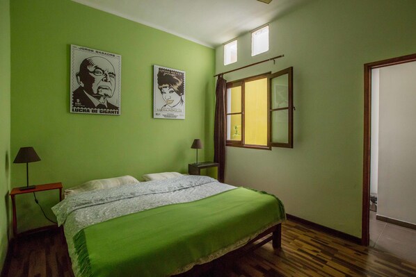 Un dormitorio con una pared verde, una cama con cubrecama verde, dos carteles, una ventana y una puerta.
