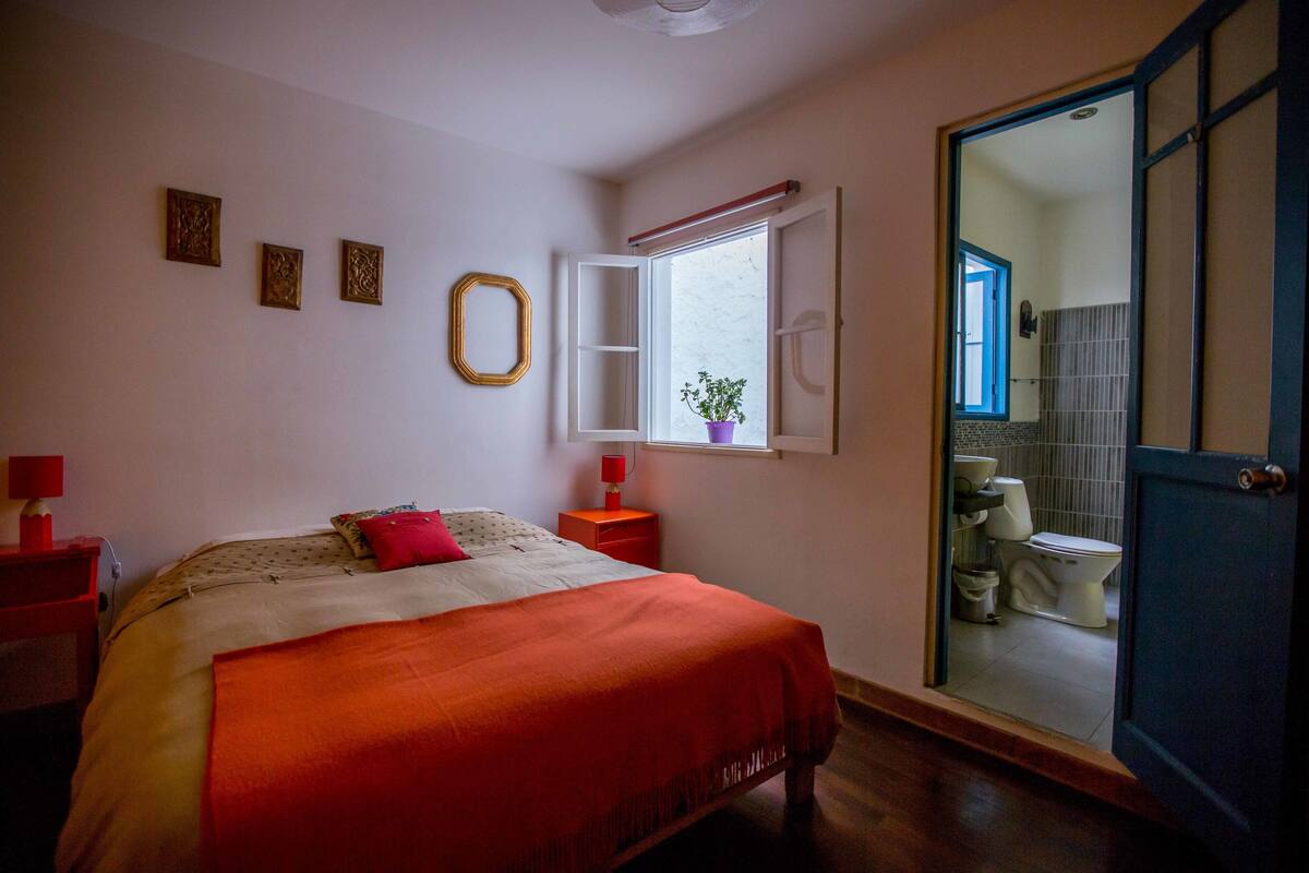 Un dormitorio con cama, mesita de noche, ventana y vistas al baño.