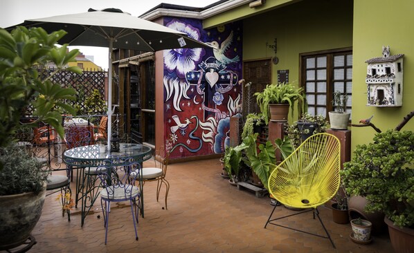 Un patio con una mesa y sillas de vidrio, rodeado de plantas en macetas y un mural.