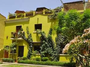 A yellow building with multiple balconies, some with plants. There are windows and a door visible.
